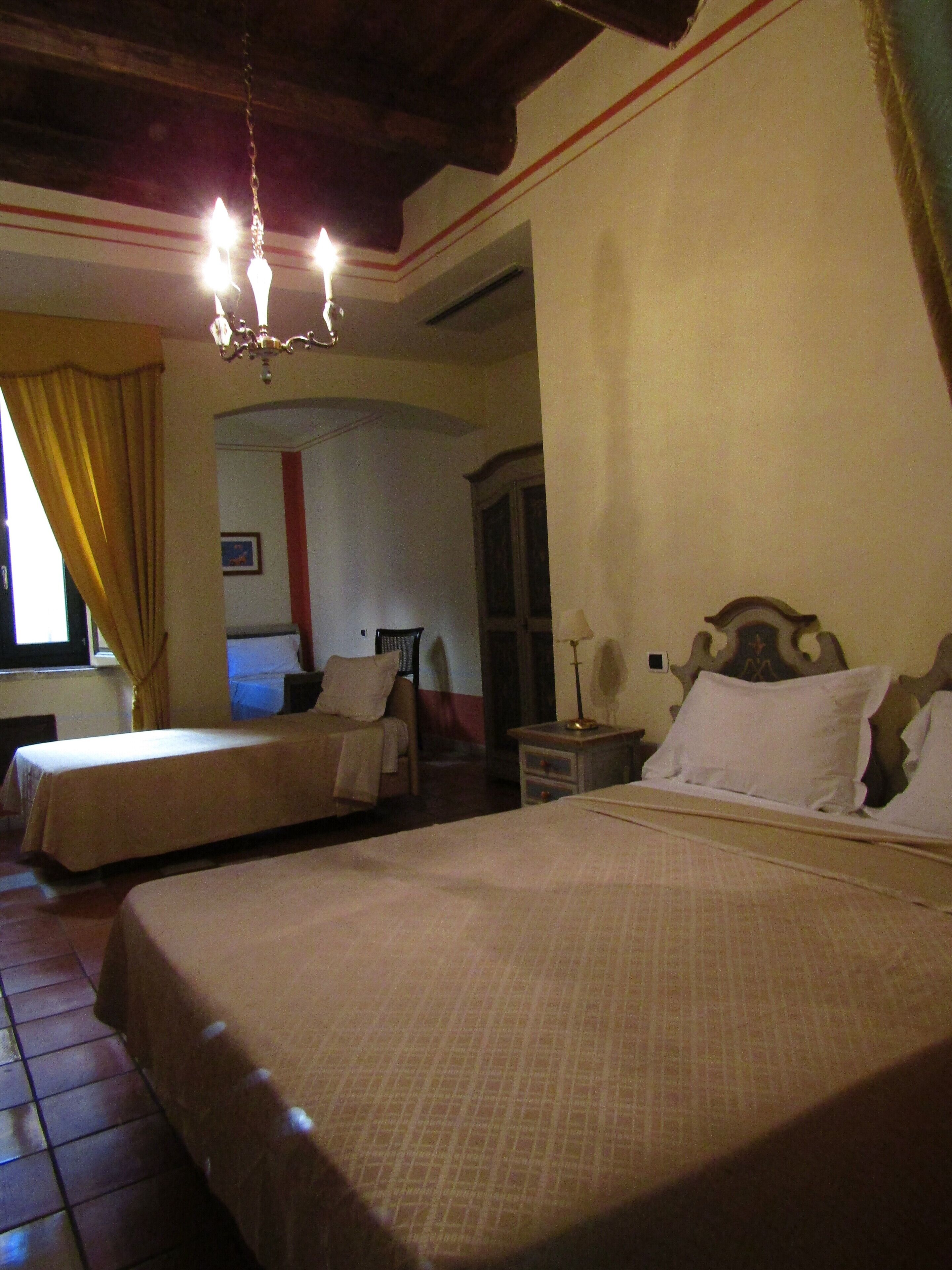 A hotel room with two beds, a chandelier, a window with curtains, and a small table with a lamp.