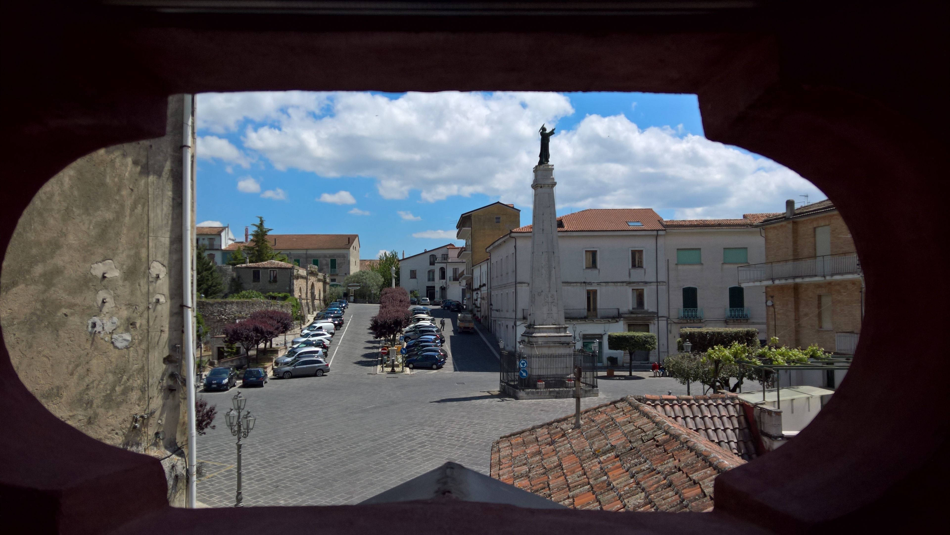Vista de la calle a través de un ventanal circular, con una estatua, coches aparcados y edificios de arquitectura tradicional.