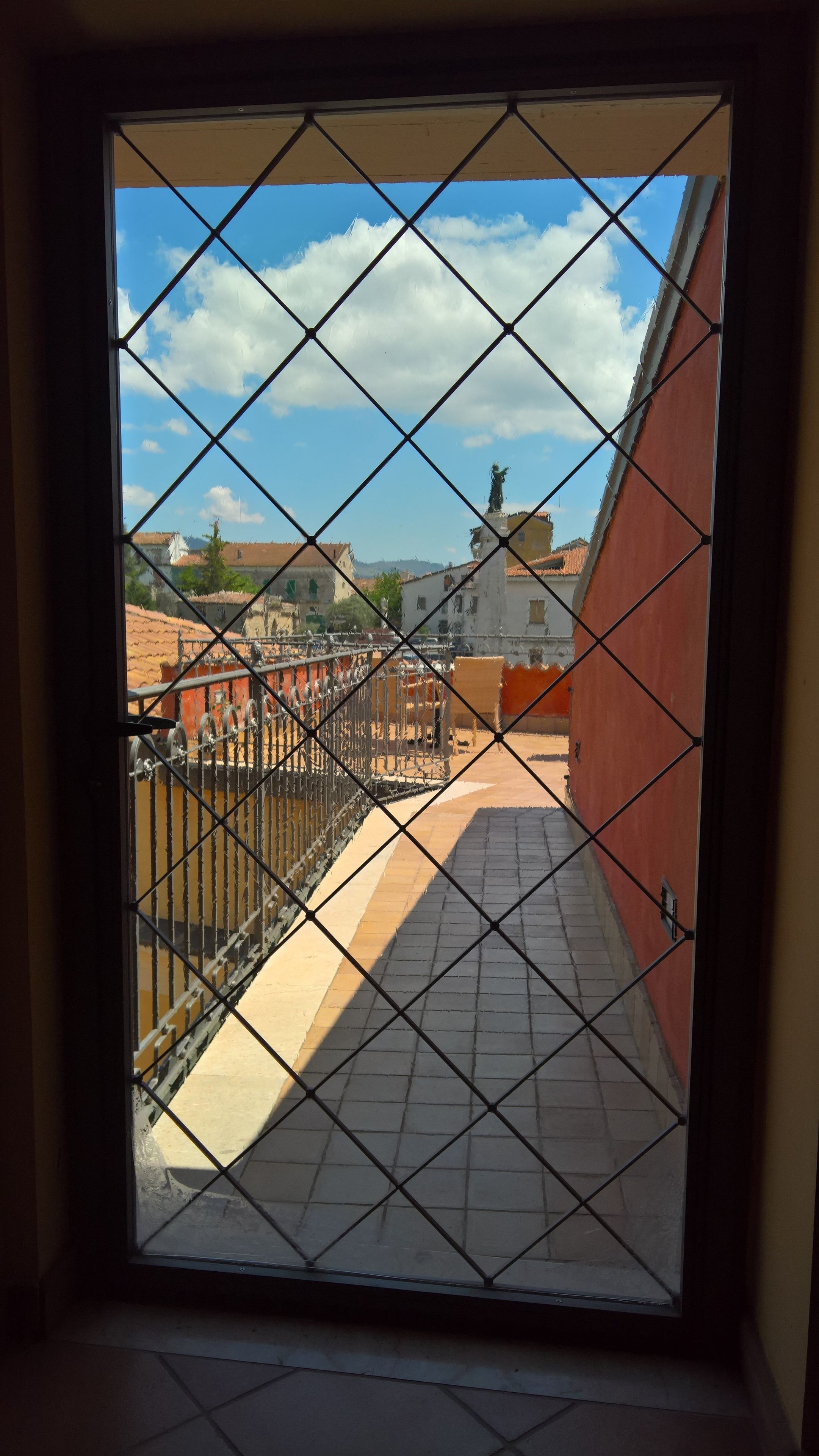 A view through a window with a metal grid, featuring a tiled floor, a red wall, and a clear sky with clouds.