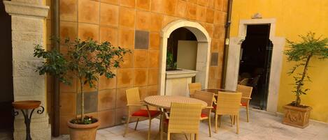 Un área de estar al aire libre con sillas de mimbre y una mesa, rodeada de plantas en macetas y un muro de baldosas.