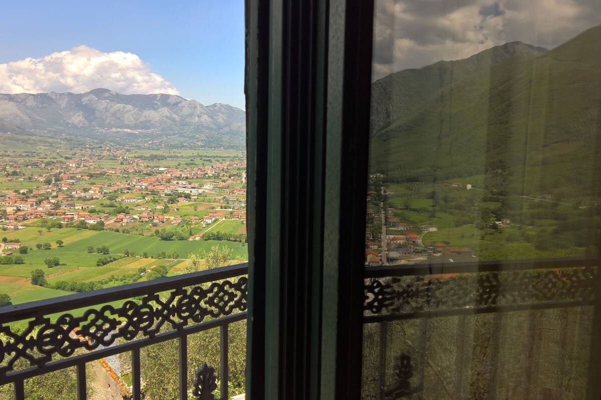 Une vue depuis un balcon doté d’une balustrade en métal, donnant sur une ville avec des bâtiments aux toits rouges et des champs verdoyants, avec des montagnes en arrière-plan.