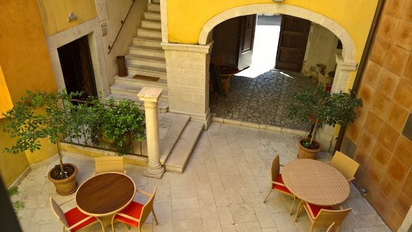 An outdoor patio area with stone flooring, two round wooden tables, wicker chairs, potted plants, and a staircase leading to an upper level.