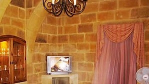 Une pièce aux murs de pierre, avec un téléviseur posé sur un meuble, une armoire en bois, un rideau rose et un ventilateur.