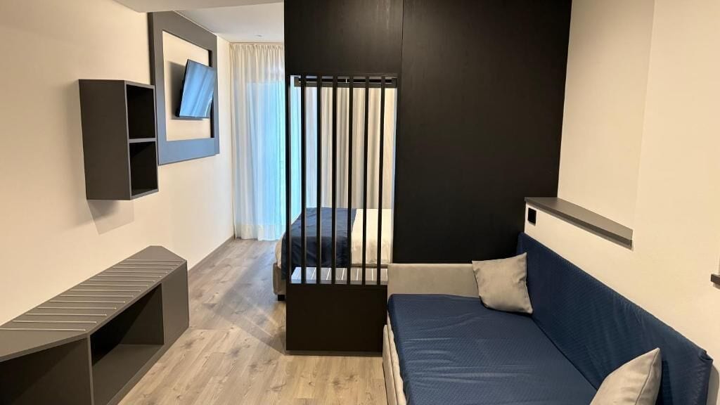 A modern hotel room with a bed, a small desk, a TV mounted on the wall, and a window with curtains.