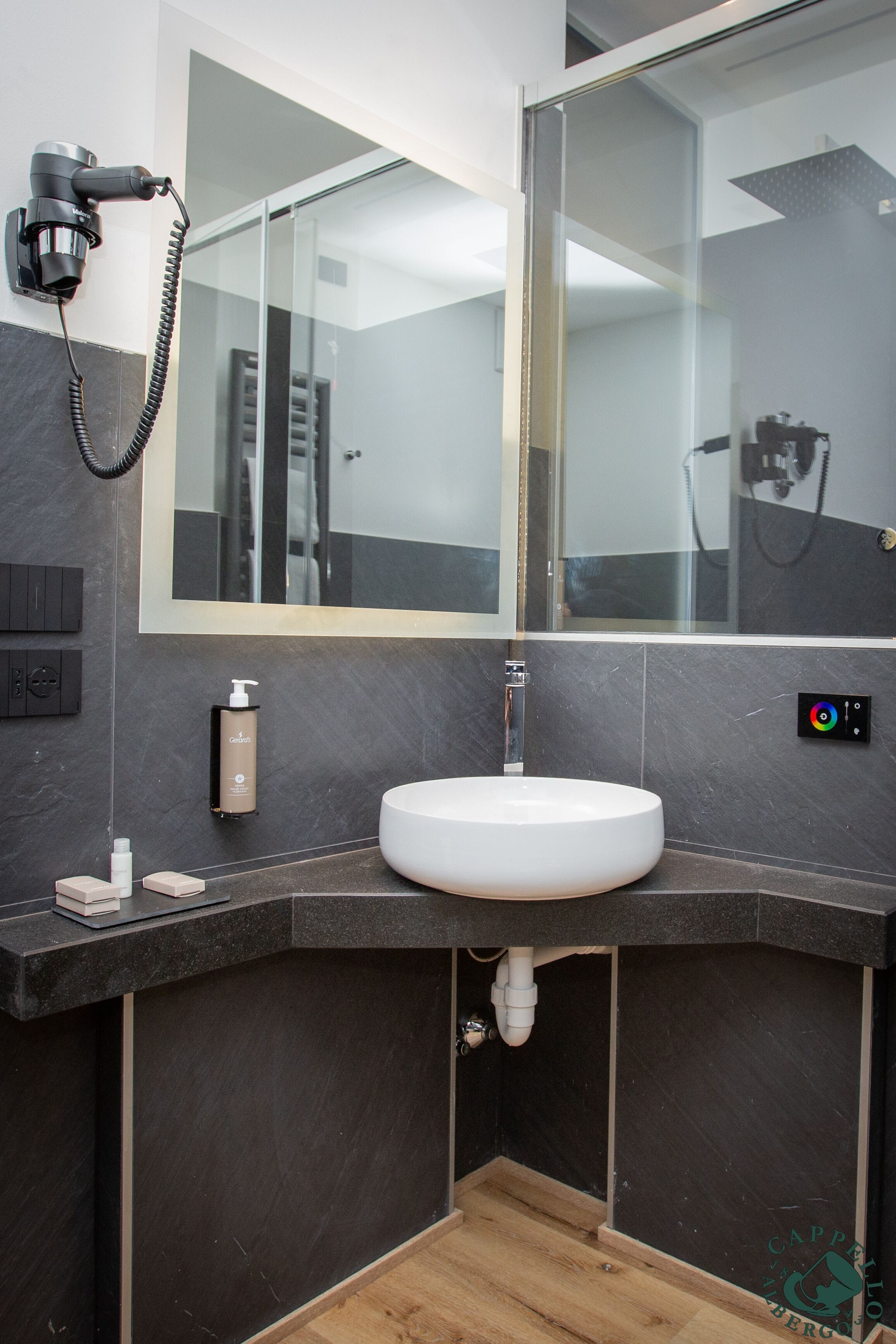 A modern bathroom with a white sink, dark tiled walls, a large mirror, and a hairdryer mounted on the wall.