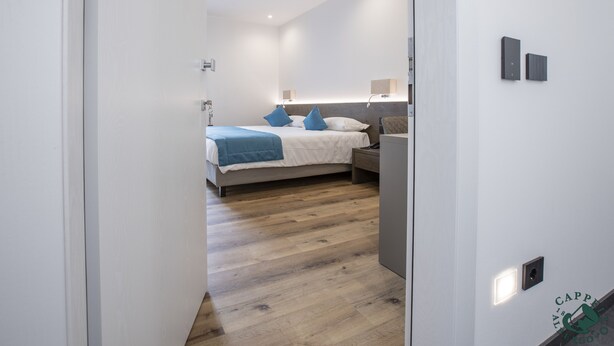 A modern hotel room with a wooden floor, a bed with blue pillows, a nightstand, and a wall-mounted lamp. - Albergo Cappello e Cadore (Belluno)