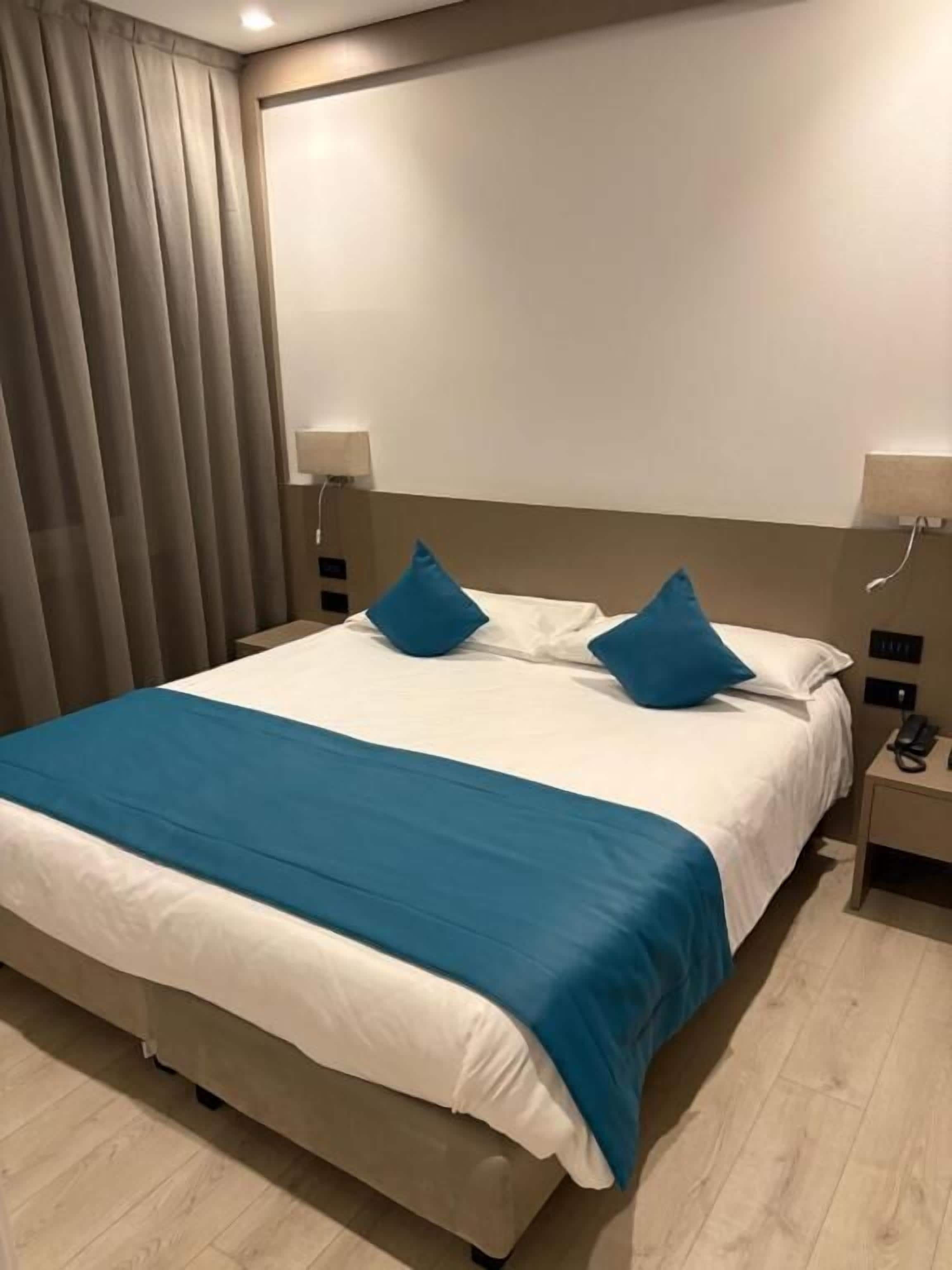 A hotel room with a large bed, two bedside tables, and a curtain on the left.