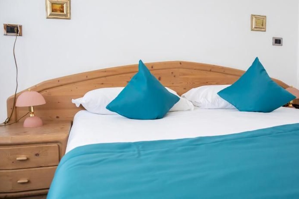 A neatly made bed with a teal bedspread and two blue pillows, a wooden headboard, a bedside lamp, and a wooden nightstand with drawers.