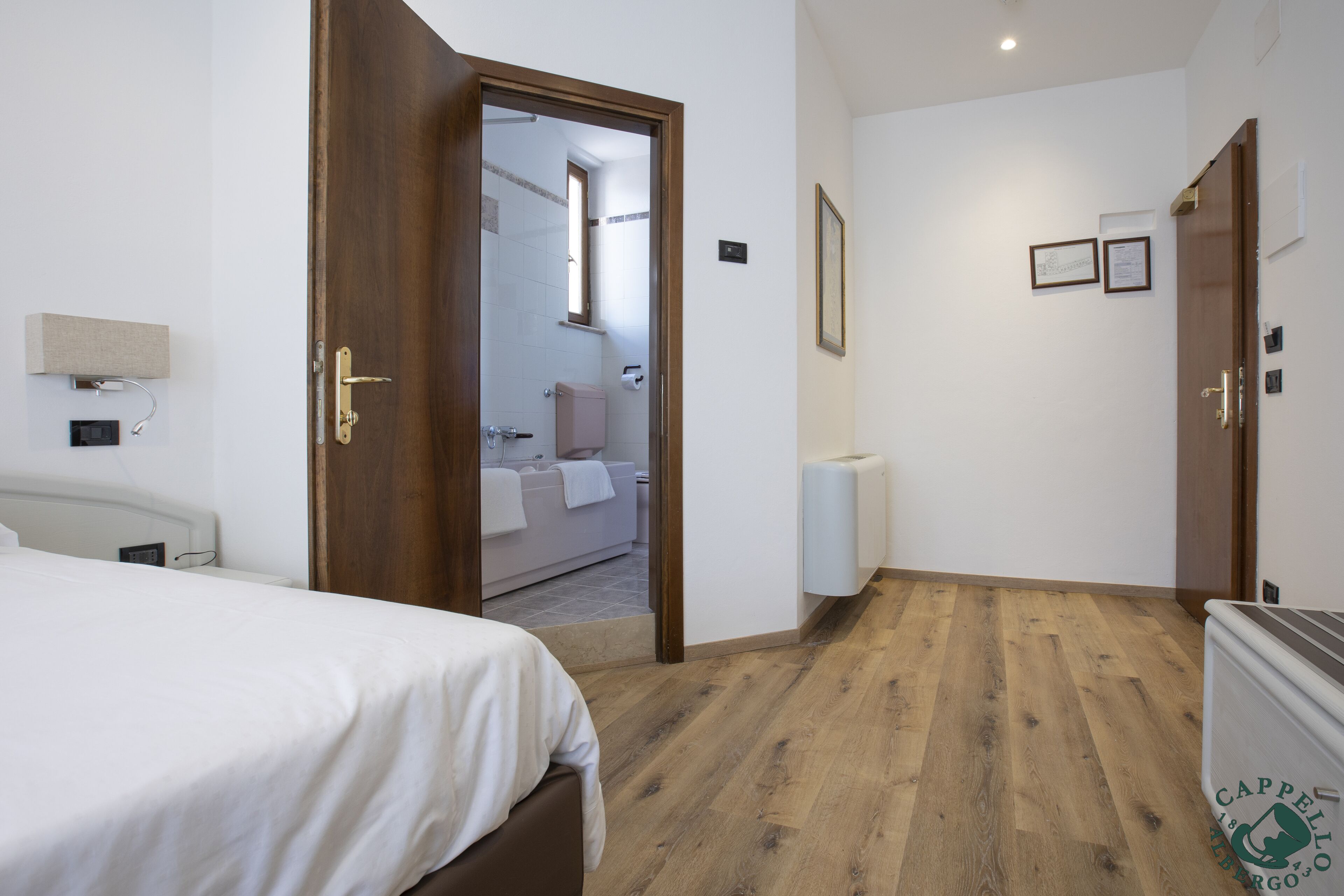 Habitación de hotel con piso de madera, una cama con ropa blanca y una puerta que da a un baño, además de un radiador.