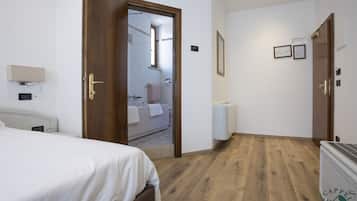Habitación de hotel con piso de madera, una cama con ropa blanca y una puerta que da a un baño, además de un radiador.