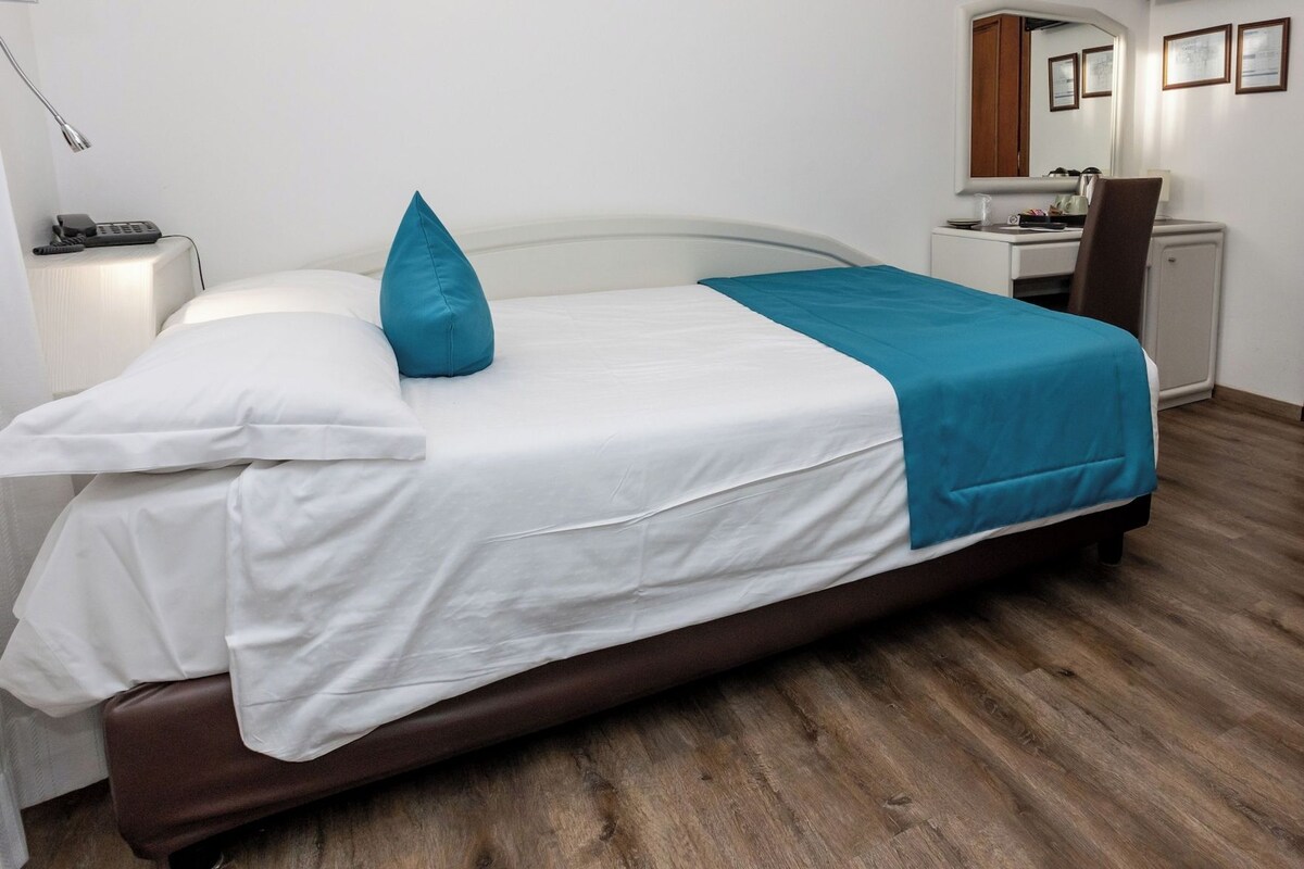 A neatly made bed with white and blue bedding, a bedside table with a lamp, and a small desk with a chair.