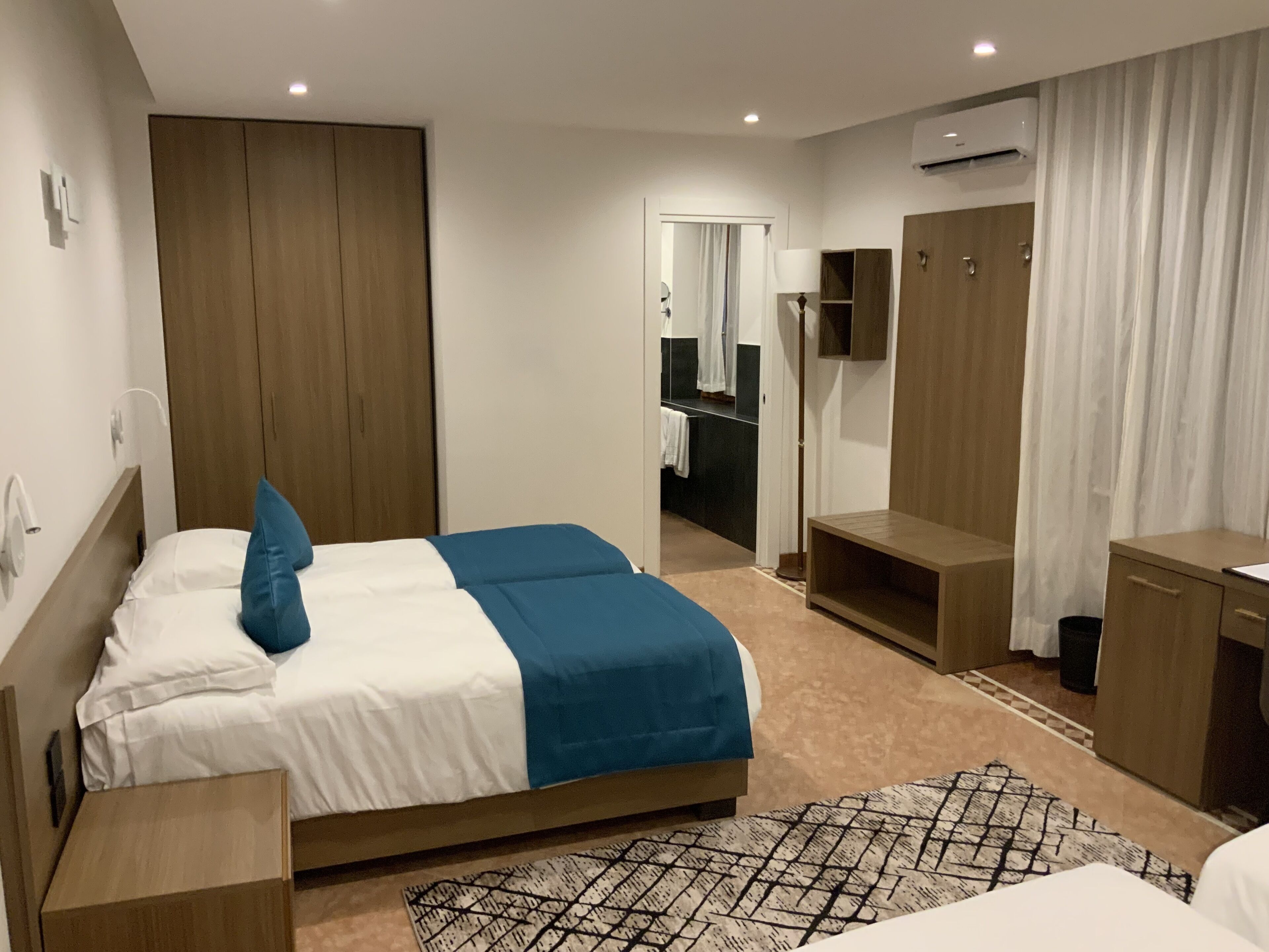 A modern hotel room with a large bed, a desk, a wardrobe, and a bathroom visible through an open door.