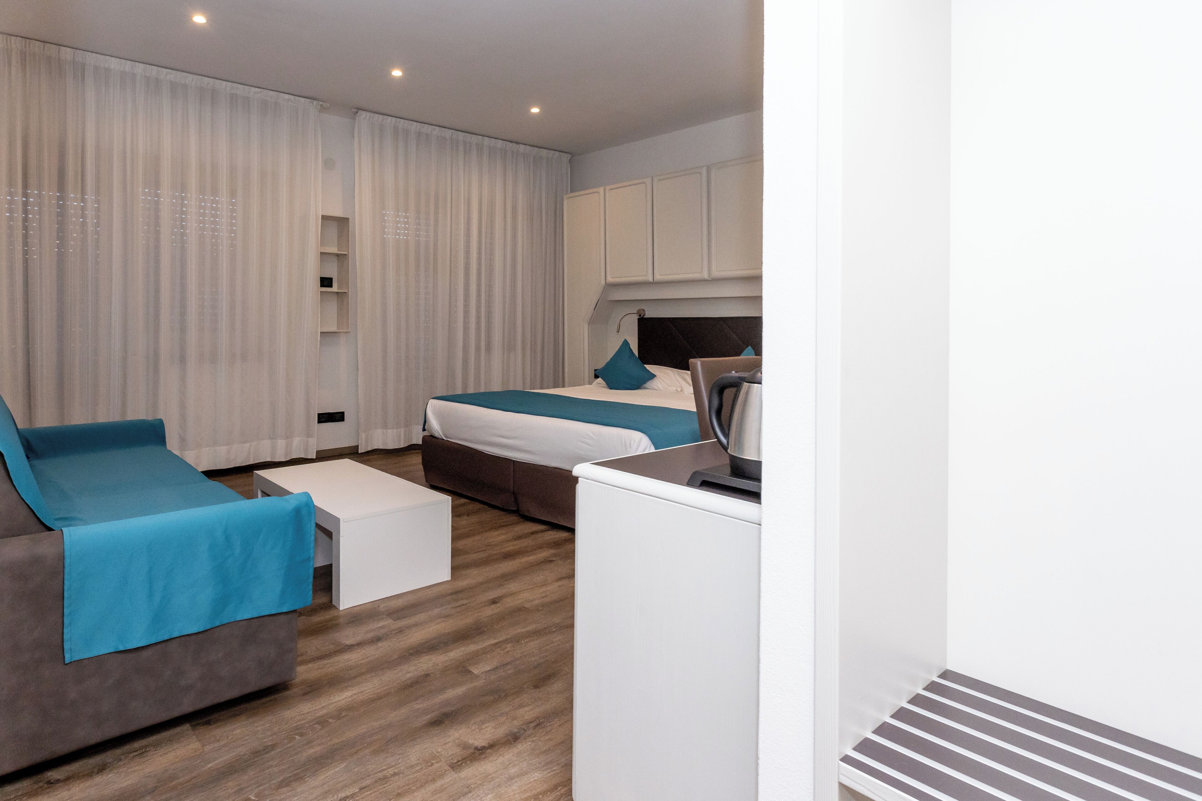Una habitación de hotel moderna con un sofá, una cama con ropa de cama azul, una mesita de centro y una zona de kitchenette.