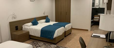 Habitación de hotel moderna con cama doble, escritorio con lámpara, armario y baño visible a través de una puerta abierta.