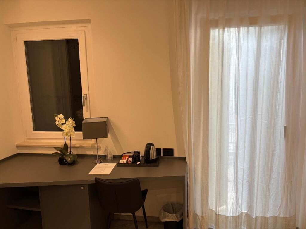 A hotel room with a desk, a chair, a window with curtains, a vase with flowers, and a lamp.