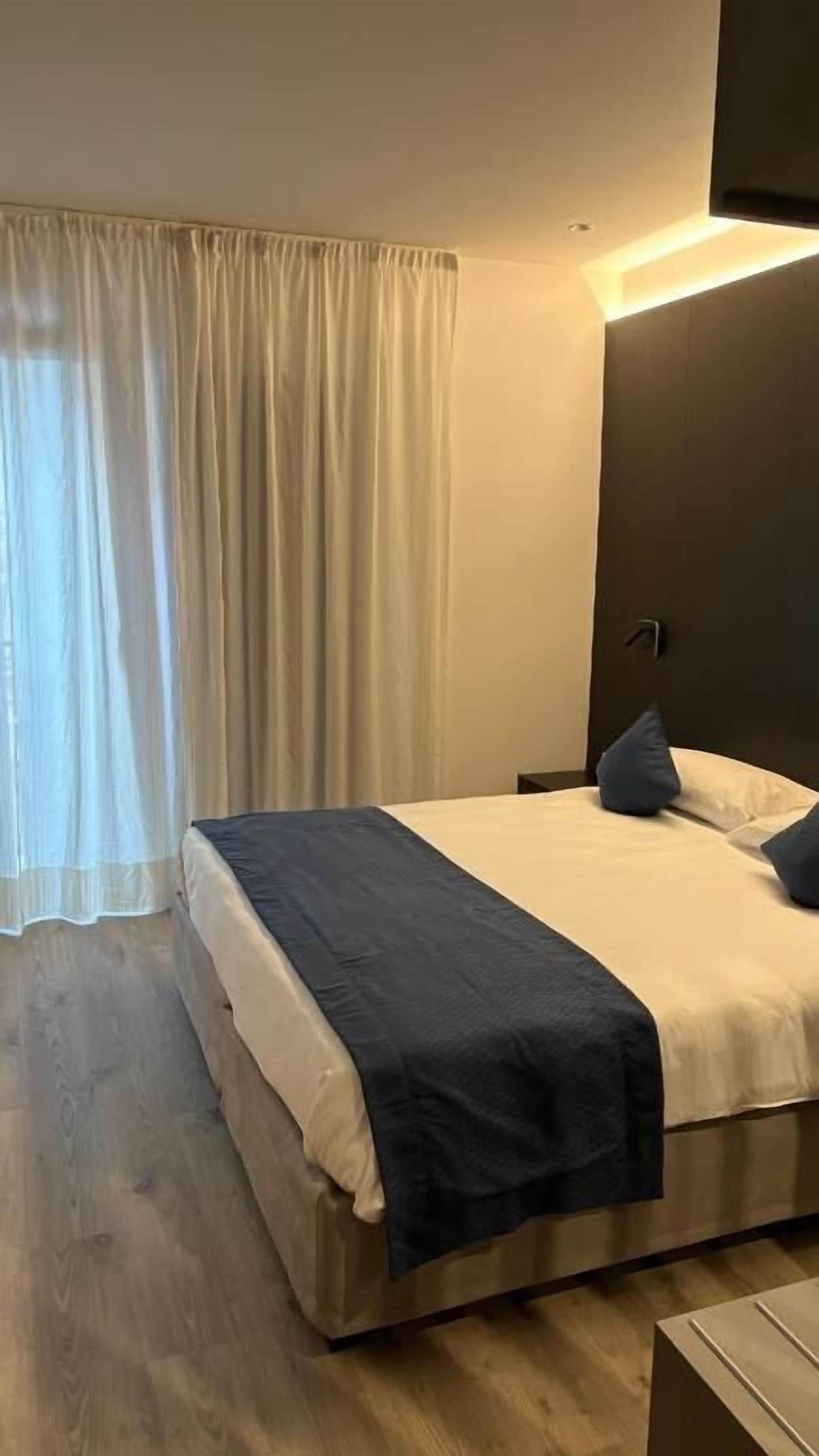 Una habitación de hotel con una cama, una mesita de noche, una lámpara y un ventanal con cortinas.