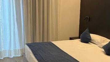 Una habitación de hotel con una cama, una mesita de noche, una lámpara y un ventanal con cortinas.