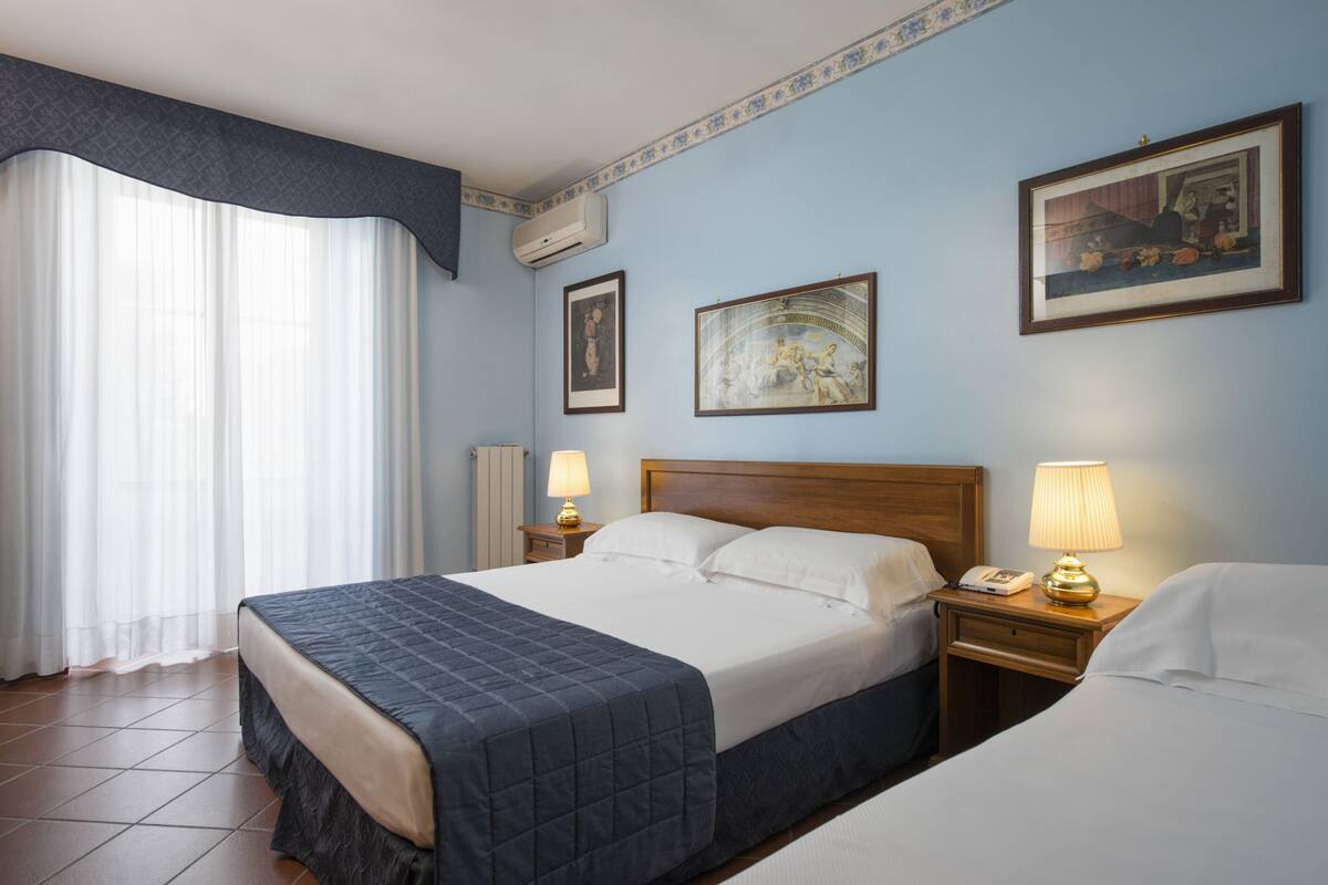 Habitación de hotel con una cama grande, dos mesitas de noche con lámparas, una ventana con cortinas translúcidas y dos cuadros en la pared.