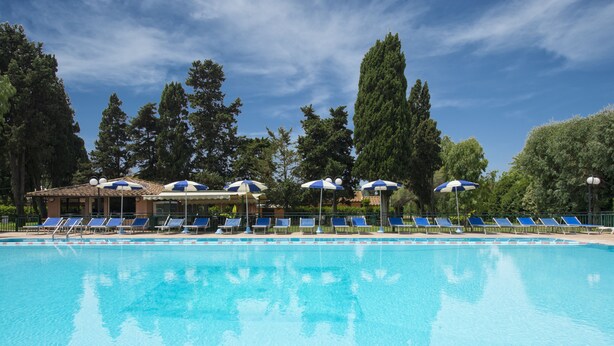 A swimming pool with blue lounge chairs and umbrellas, surrounded by trees and a building. - La Pineta dei Liberti (Ardea)