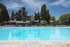 Una piscina con sillas reclinables azules y sombrillas, rodeada de árboles y un edificio.