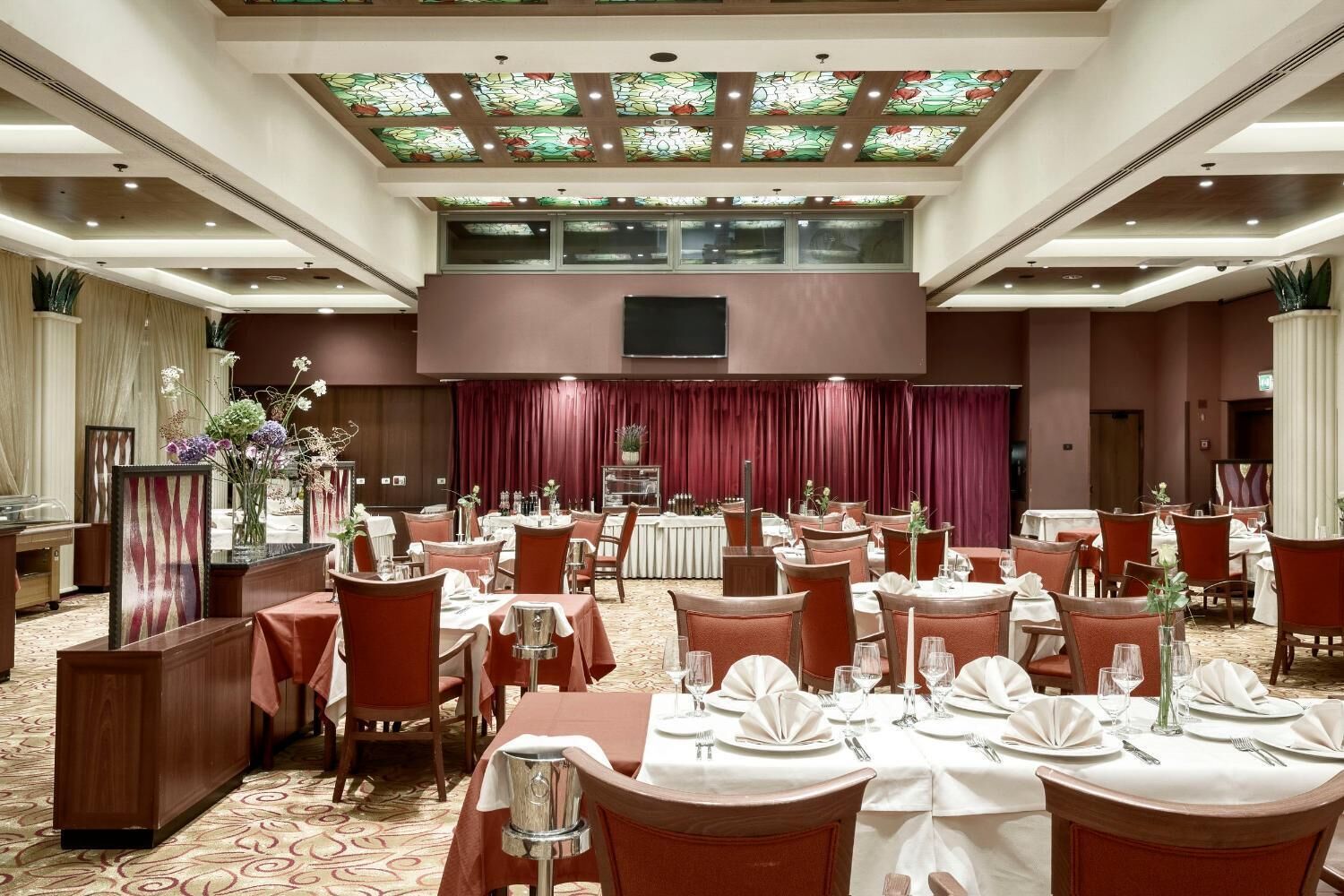 Zona de menjador d'hotel amb diverses taules preparades per a comensals, un escenari amb cortines i un sostre decoratiu.