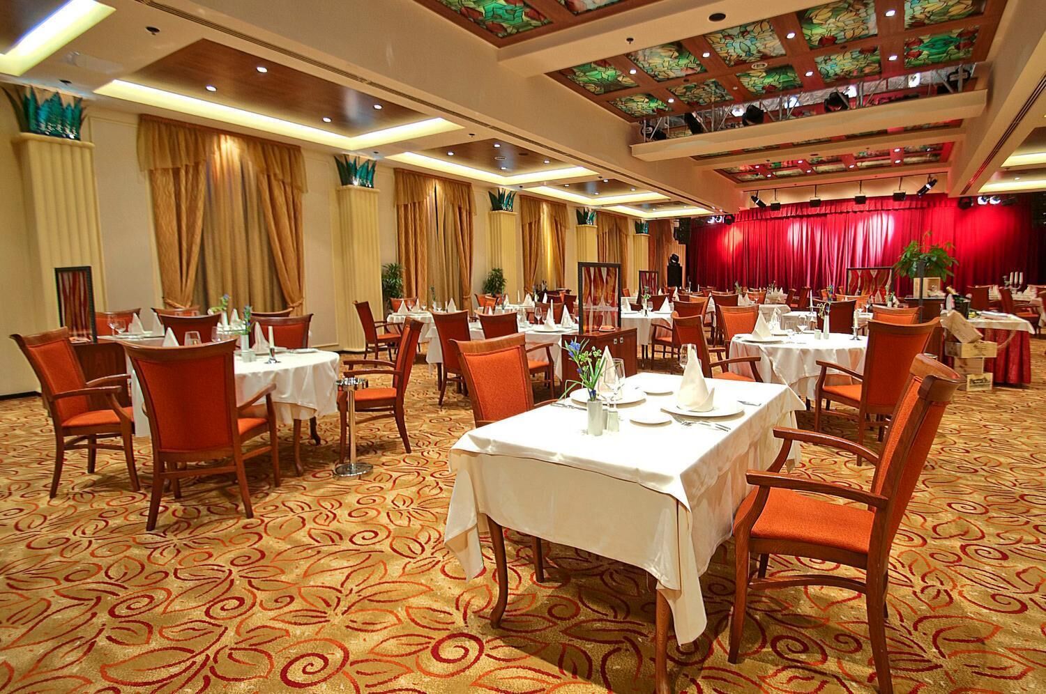 A banquet hall with round tables, chairs, and a stage with red curtains.