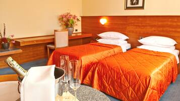 Une chambre d’hôtel avec deux lits, une table de chevet sur laquelle se trouve une bouteille de champagne et des verres, une petite plante en pot et une lampe fixée au mur.