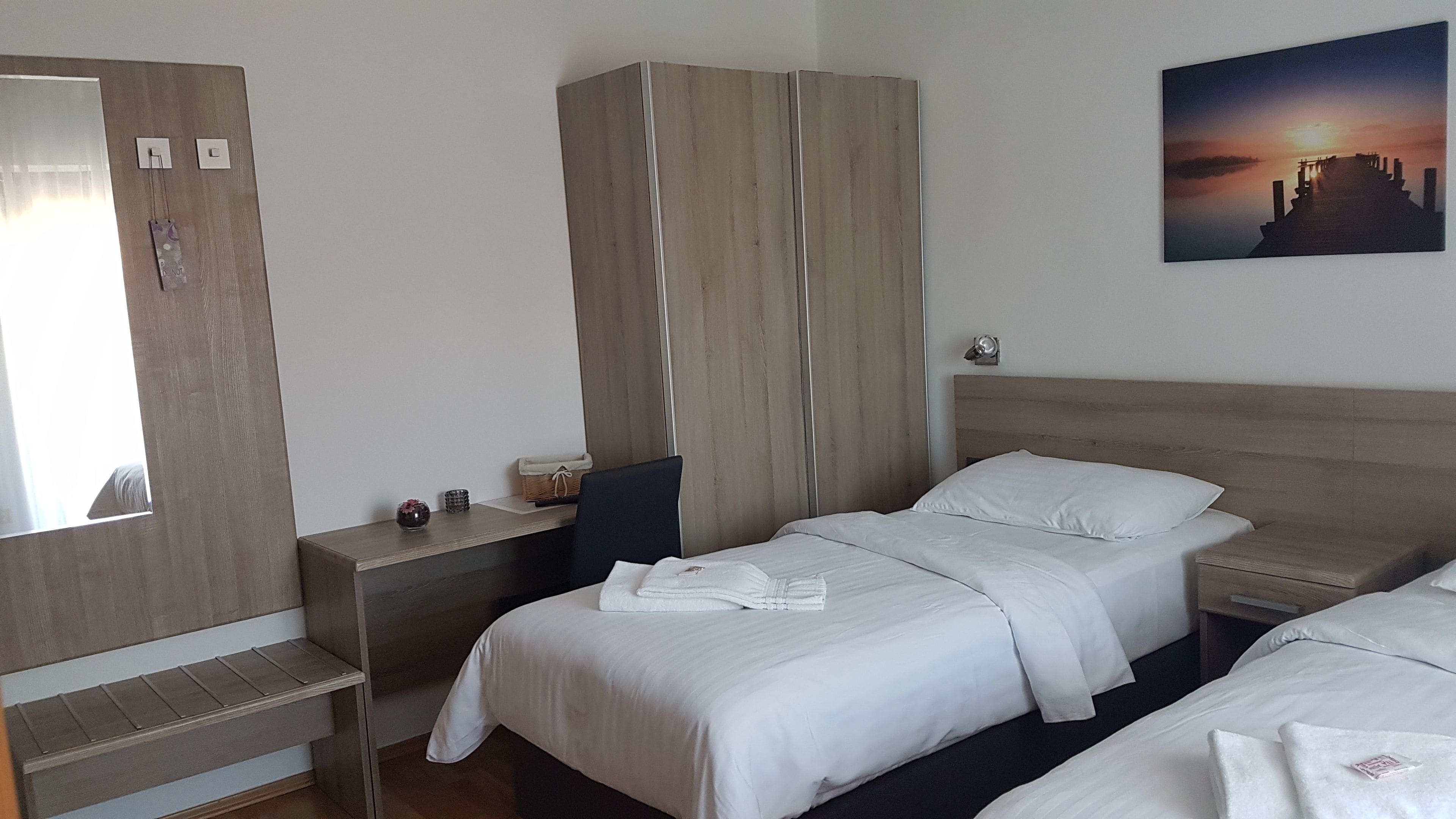 A hotel room with two beds, a wardrobe, a desk, a chair, a nightstand, a mirror, and a painting on the wall.