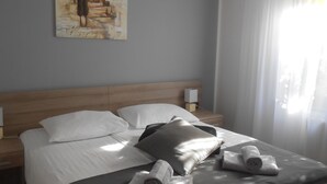 Un lit bien fait, avec du linge de lit blanc, deux oreillers et deux serviettes enroulées. On y trouve une tête de lit en bois, une table de chevet avec une lampe et un tableau accroché au mur.