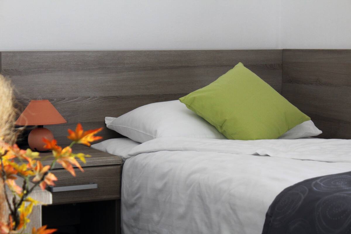 A neatly made bed with white bedding and a green pillow, a wooden nightstand with a lamp, and a vase with flowers.