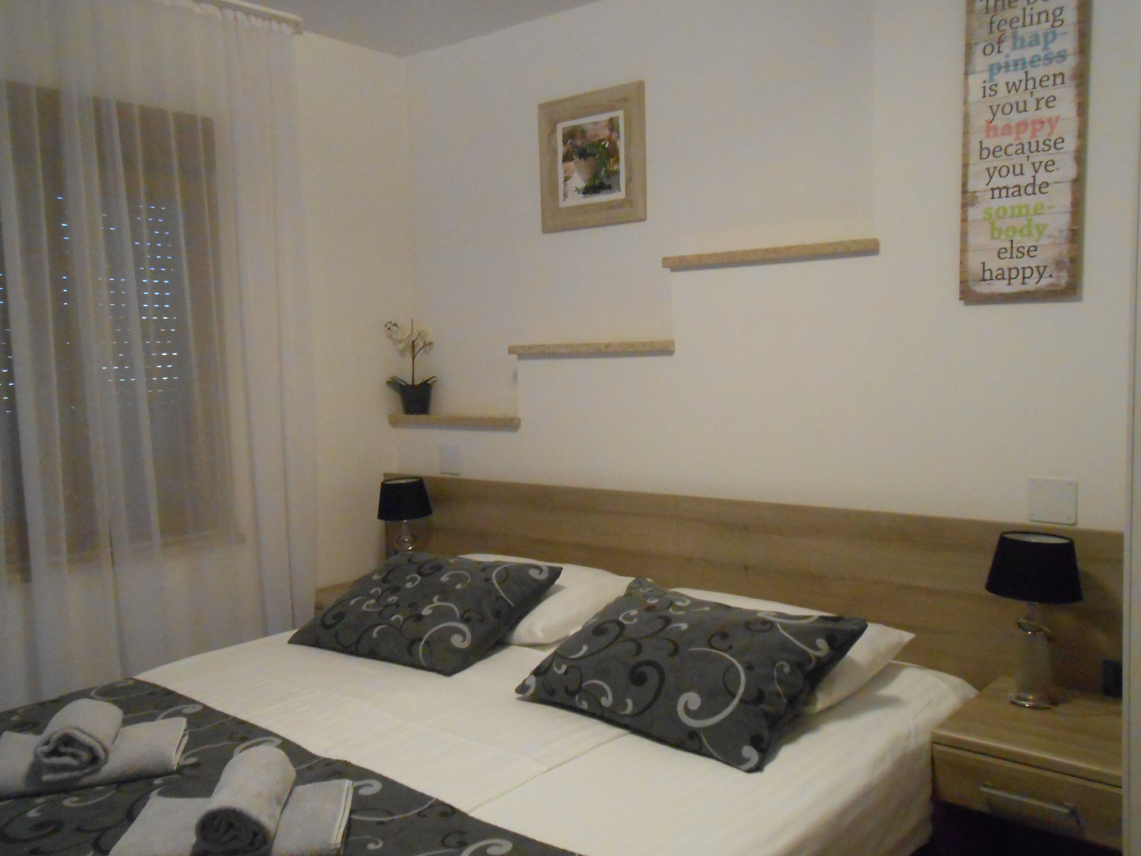 A neatly made bed with patterned pillows, a bedside table with a lamp, a small potted plant, and a framed picture on the wall.