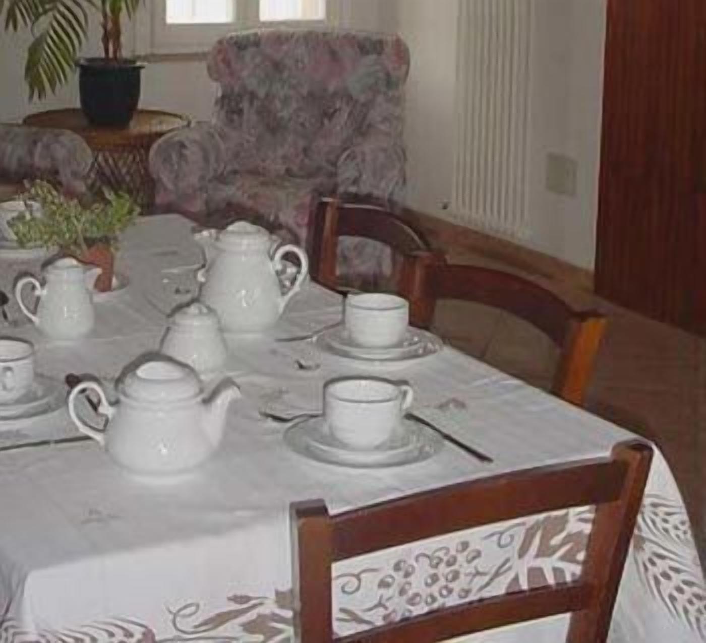 Une table à manger est dressée avec des théières, des tasses et des soucoupes blanches, et entourée de chaises en bois recouvertes d’un tissu à motifs floraux.