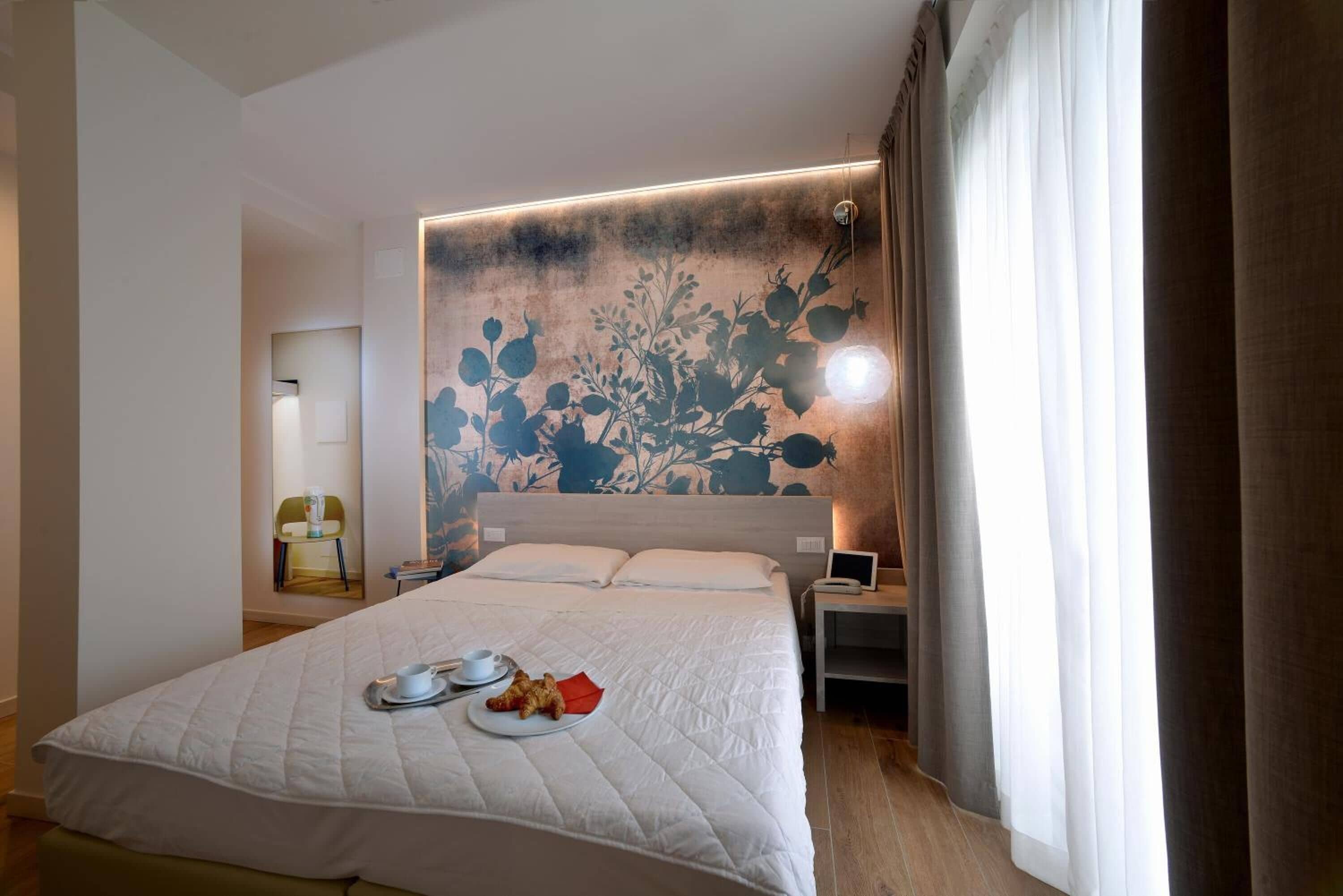 Ein Hotelzimmer mit einem großen Bett, einem Nachttisch mit Lampe, einem an der Wand befestigten Kunstwerk und einem Fenster mit Vorhängen.