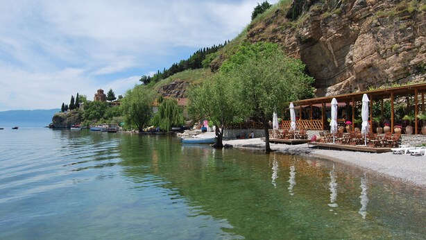 A lakeside resort with clear water, a wooden deck with umbrellas, and a rocky hillside in the background. - Apartments Kosta (Ohrid)
