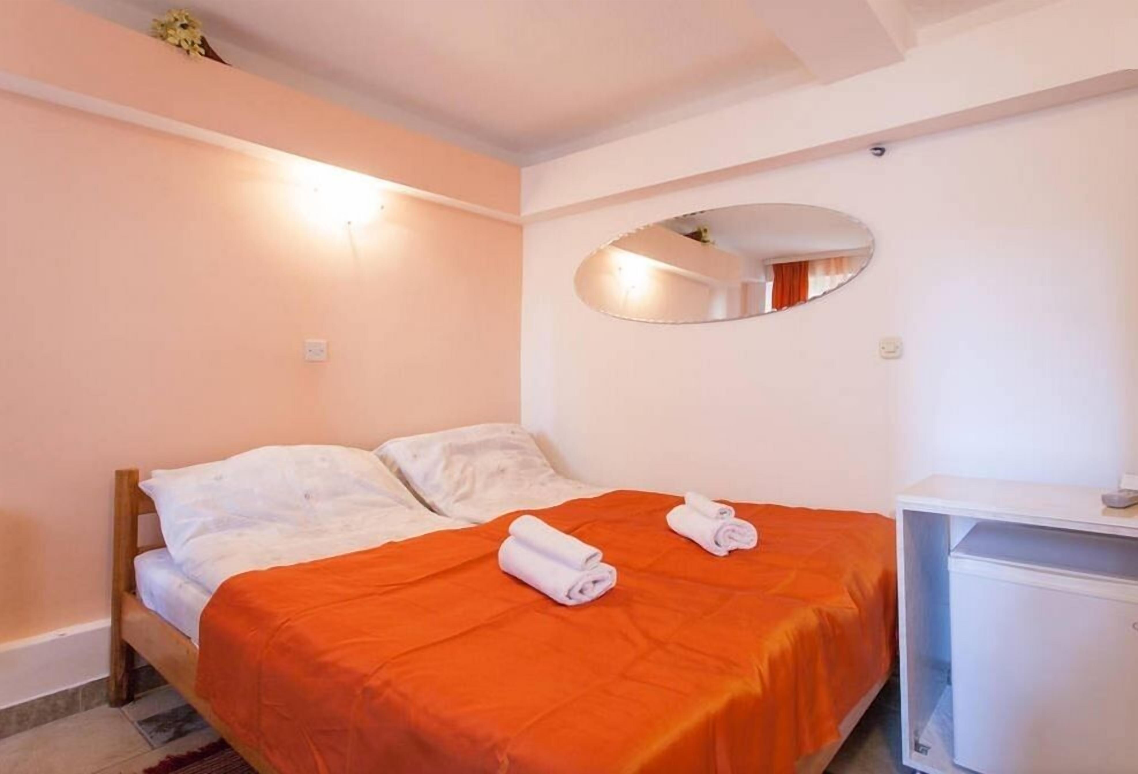 Een slaapkamer met een bed, een oranje dekbedovertrek, twee opgerolde handdoeken, een witte nachtkastje en een spiegel aan de muur.