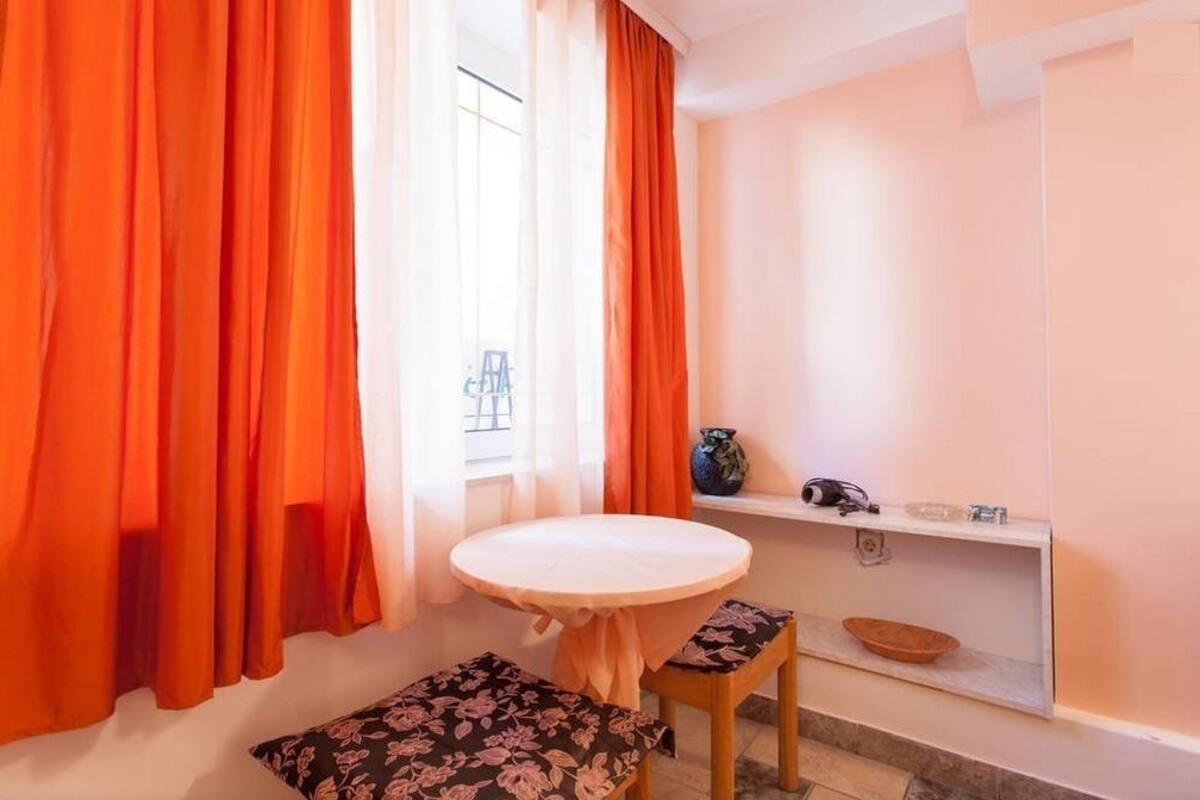 A room with a round table, two floral cushions, a shelf with a vase, and orange curtains.