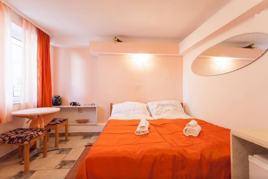 A hotel room with a bed, a small table with a chair, a desk, and a mirror.