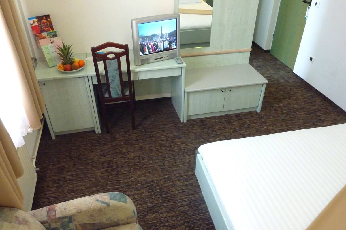 Habitación de hotel compacta con cama, un escritorio pequeño con televisor, una silla y una mesita con frutas.