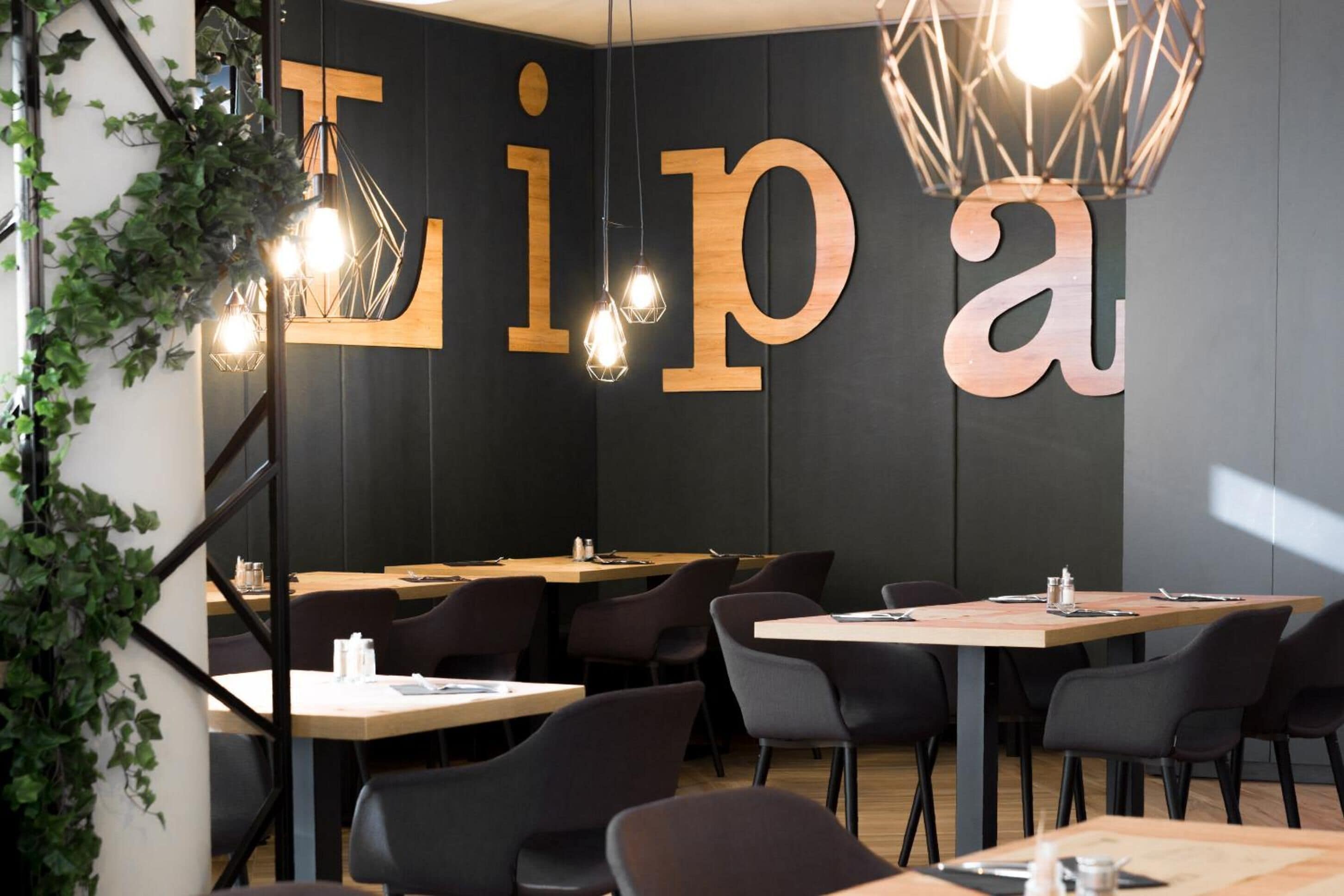 Een modern restaurantinterieur met een groot 'Lip a' bord, houten tafels, zwarte stoelen en hangende pendelverlichting.