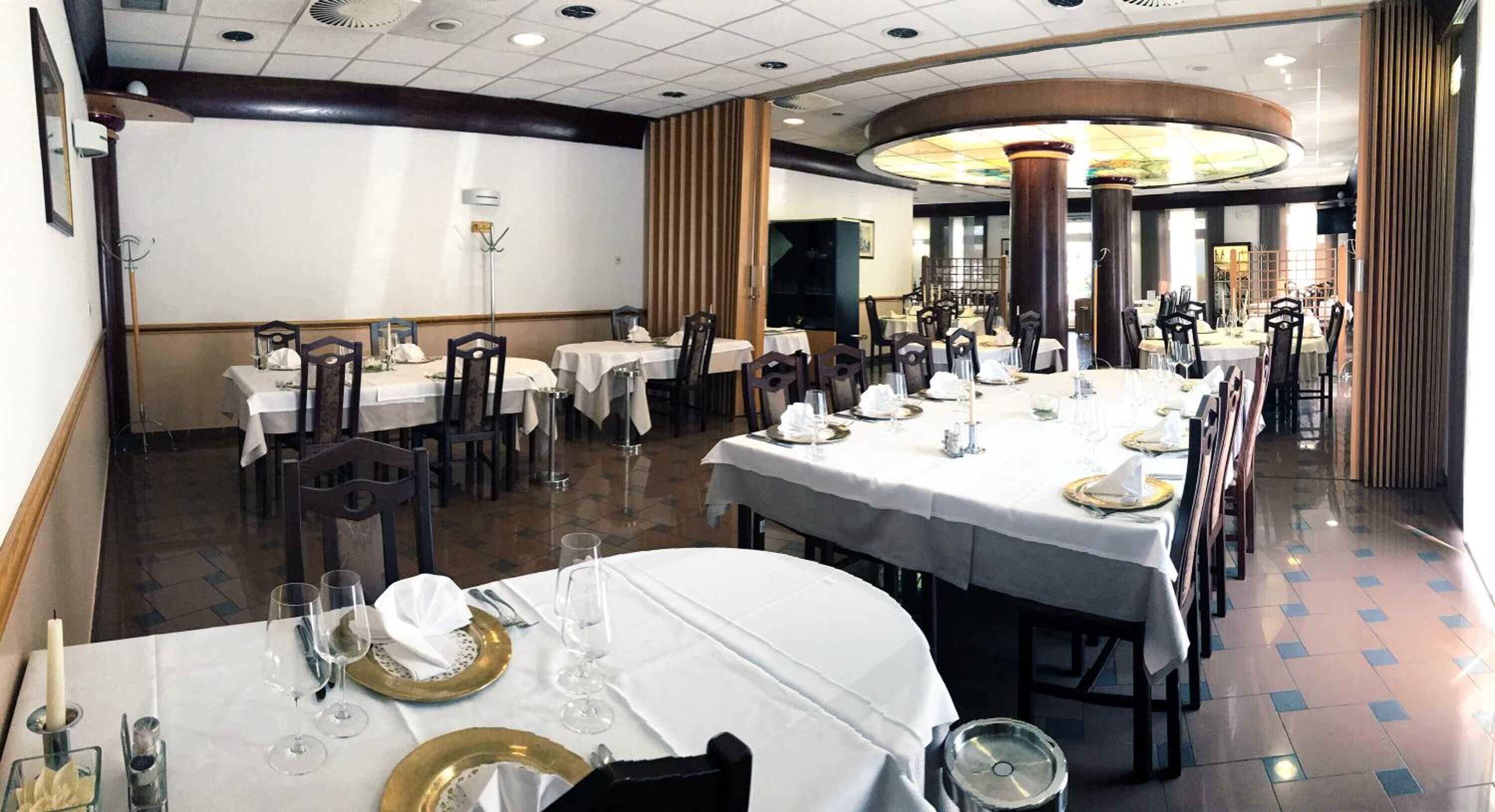 Un restaurante con varias mesas redondas dispuestas para comer, con manteles blancos y platos con detalles dorados.