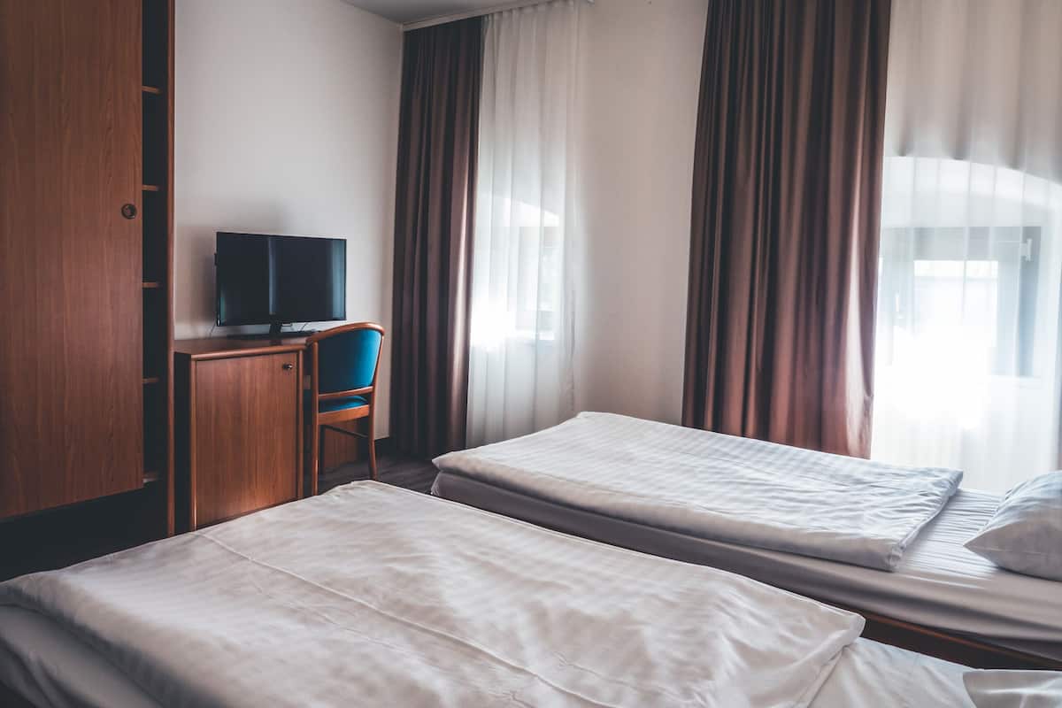 Ein Hotelzimmer mit zwei Einzelbetten, einem Holzschrank, einem Schreibtisch mit Fernseher und Vorhängen.
