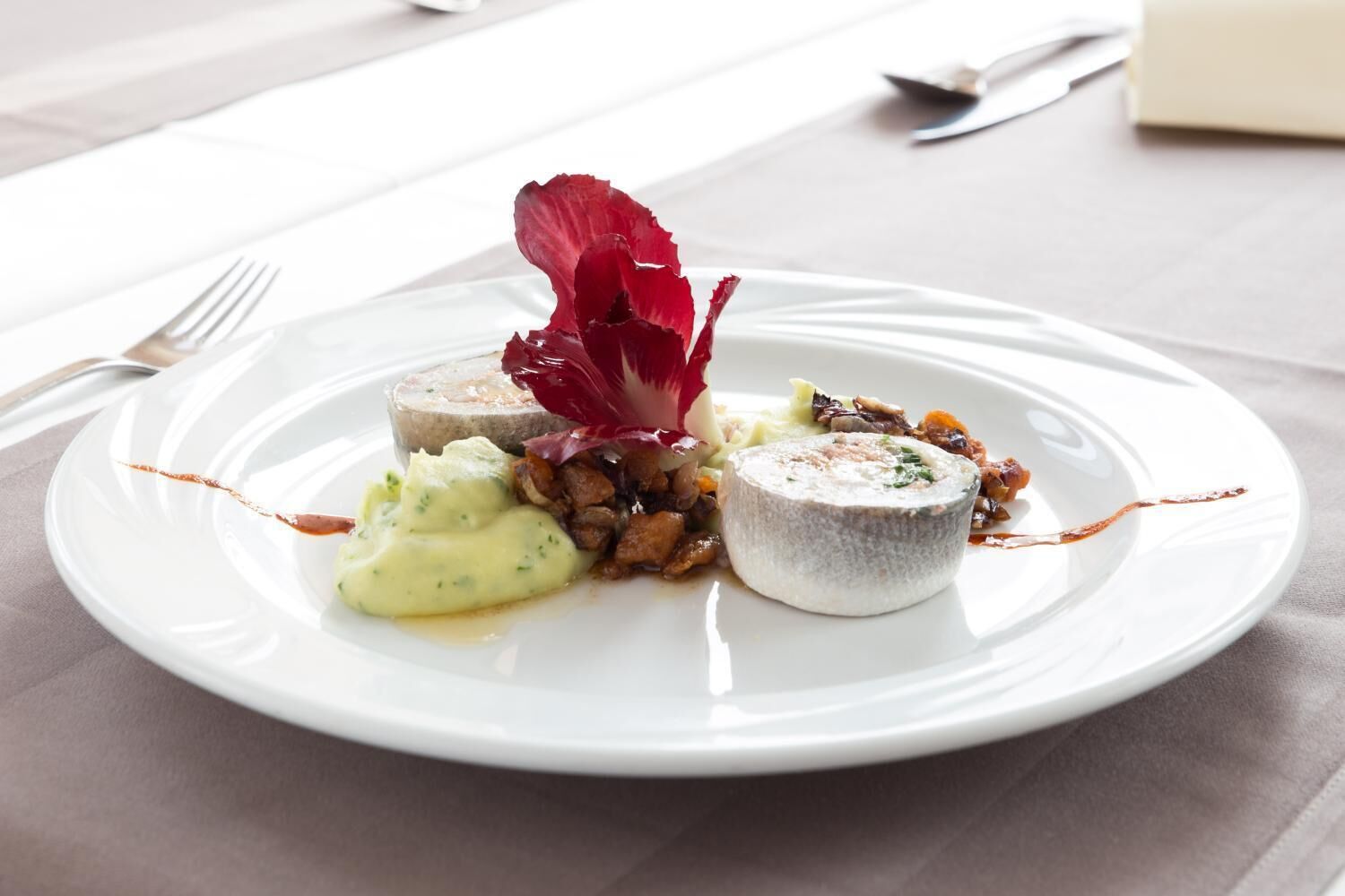 Een wit bord met diverse soorten eten, waaronder een stuk vlees, een romige saus en een klein, rond voorwerp, alles op een tafel met een bestekset op de achtergrond.