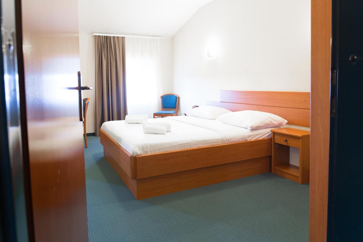 Habitación de hotel con cama de madera, una mesita de noche, una silla y vista al pasillo.