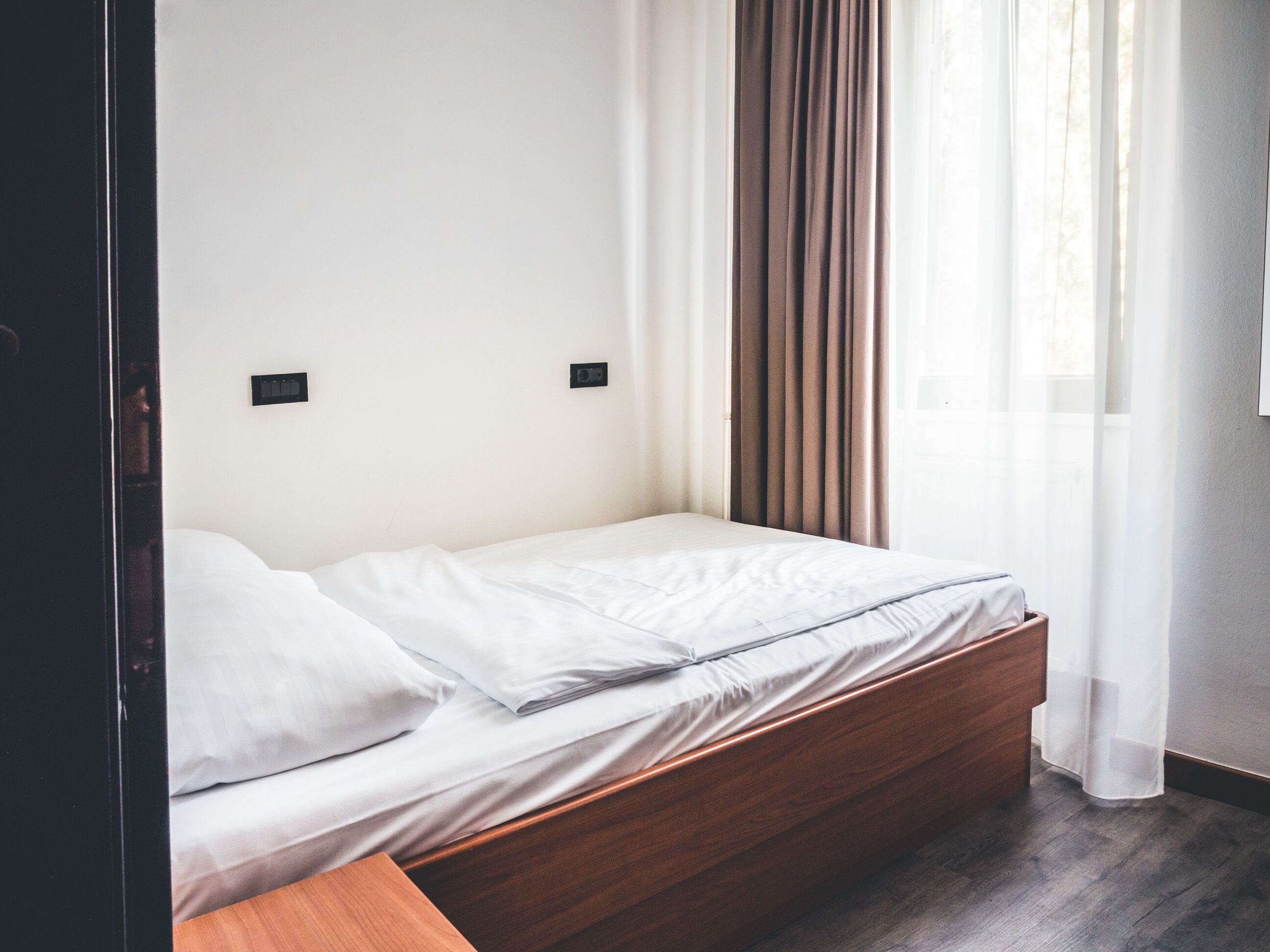 A single bed with a wooden frame, white bedding, and a window with sheer curtains.