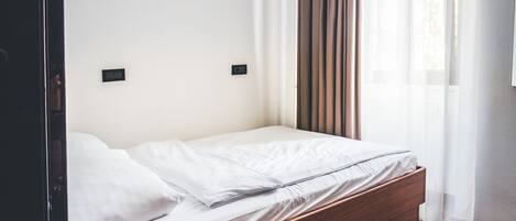 A single bed with a wooden frame, white bedding, and a window with sheer curtains.