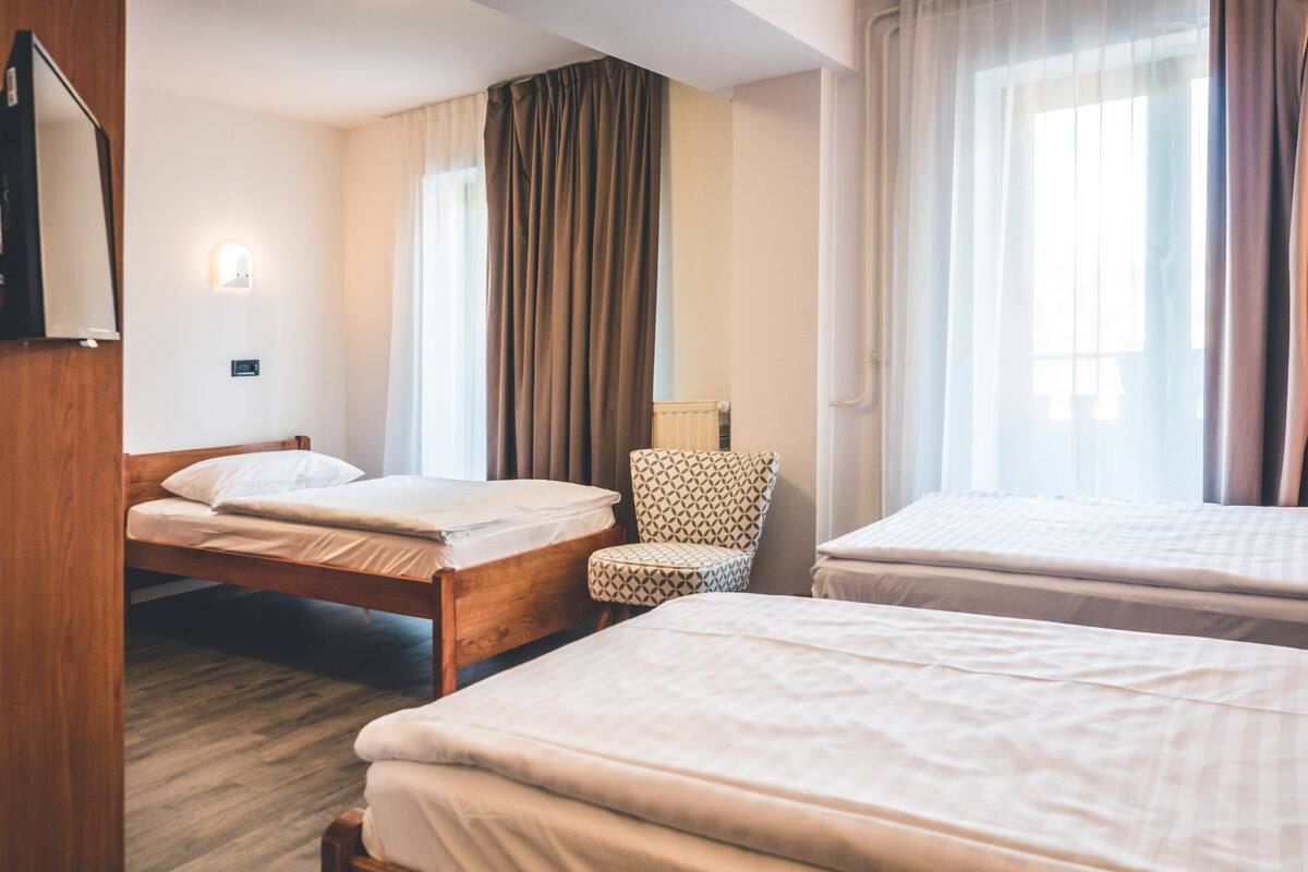 Ein Hotelzimmer mit zwei Betten, einem Sessel, einem Heizkörper und einem Fenster mit Vorhängen.
