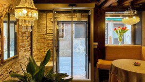 Une pièce avec un mur en pierre, des poutres en bois, un lustre, une porte coulissante et une fenêtre ornée d’un vase de fleurs.