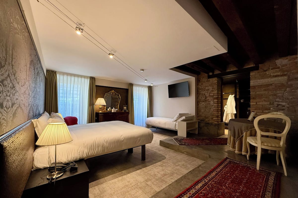 A modern hotel room with a large bed, a desk, a chair, a TV, and a fireplace.