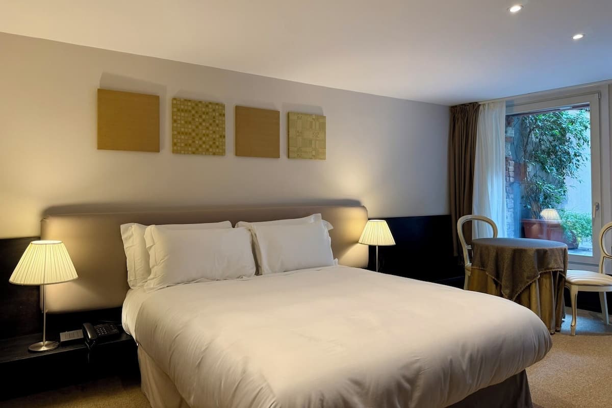 A hotel room with a bed, two bedside lamps, a small table with a chair, and a window with curtains.