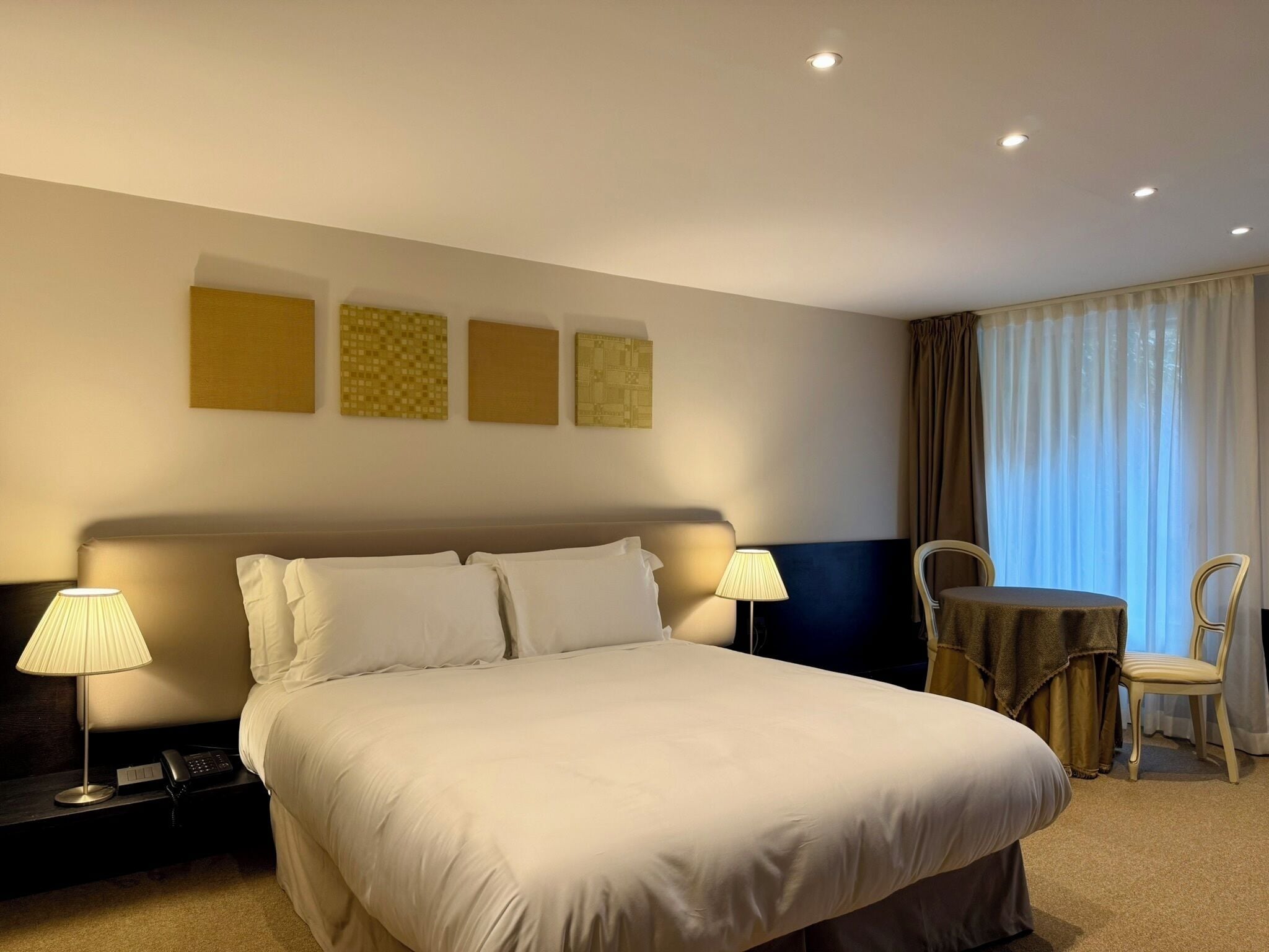 A hotel room with a large bed, bedside lamps, a desk with a telephone, and a chair at a small table.